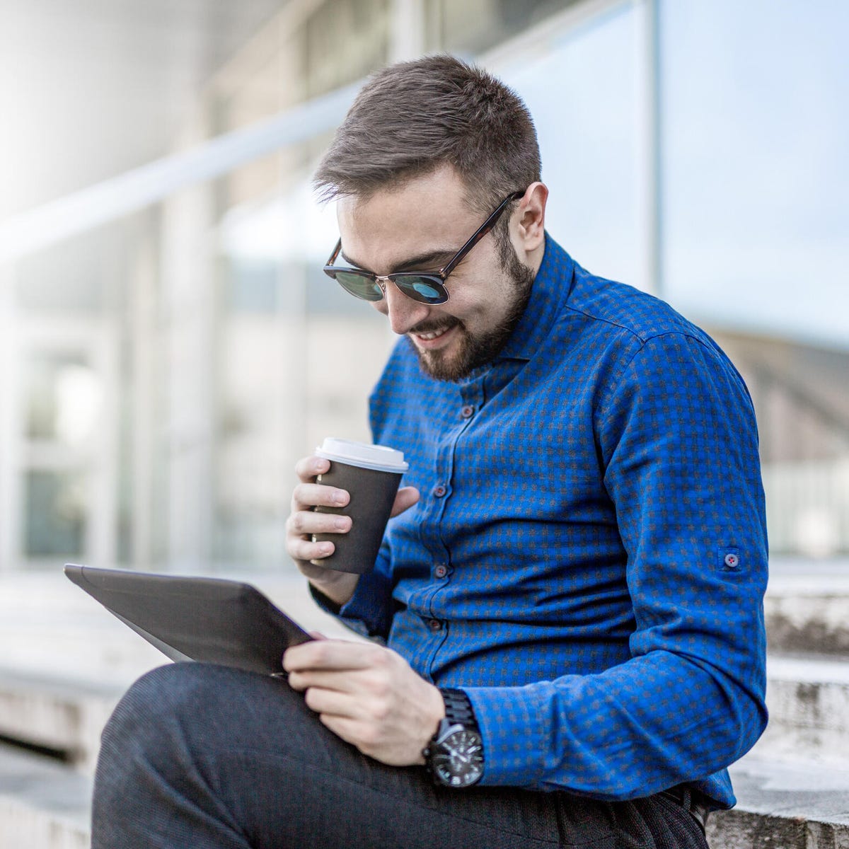 Smiling man in blue long sleeve holding a cup of coffee while looking his tablet