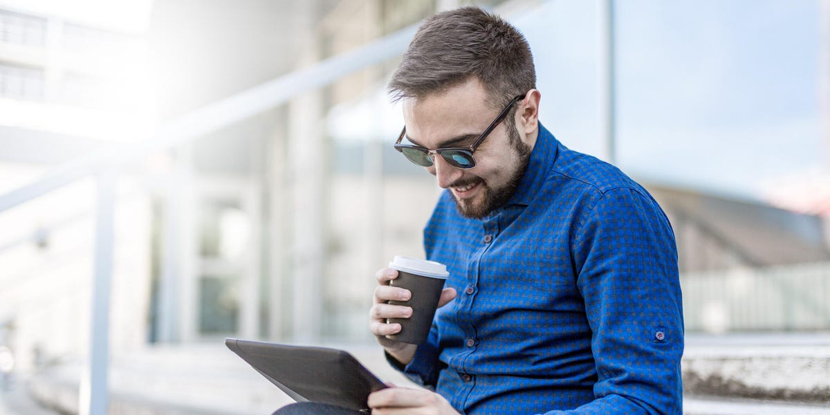 Smiling man in blue long sleeve holding a cup of coffee while looking his tablet